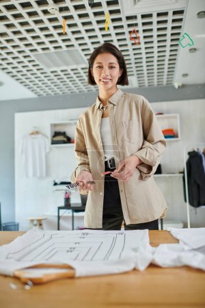 fröhliche asiatische Designerin hält Textvorlagen in der Nähe von Kleidung und blickt in die Kamera im Druckstudio