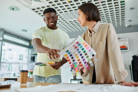 smiling african american designer pointing at color palette near asian colleague in print studio