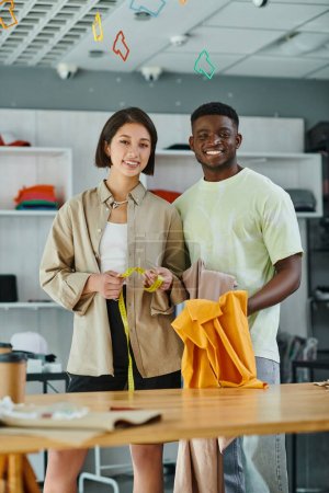 Photo for Happy interracial clothes designers with fabric and measuring tape looking at camera in print studio - Royalty Free Image