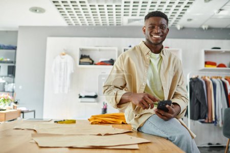 Photo for Cheerful african american designer sitting with mobile phone near sewing patterns in print studio - Royalty Free Image