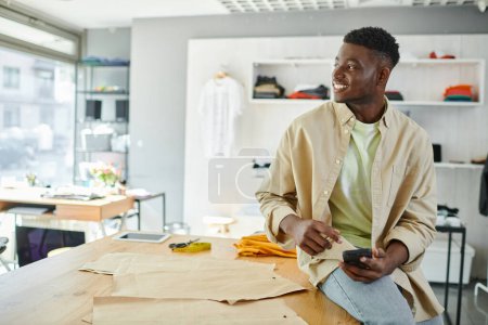 joyful african american entrepreneur sitting with smartphone and looking away near sewing patterns
