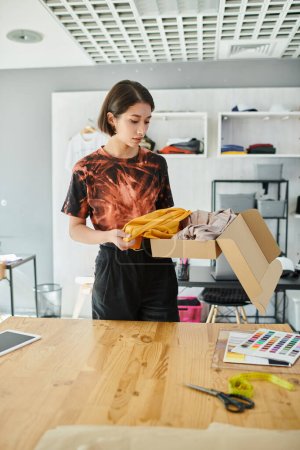 stilvolle asiatische Modedesignerin beim Auspacken von Kleidung aus Karton in der Nähe von Farbmustern im Druckstudio