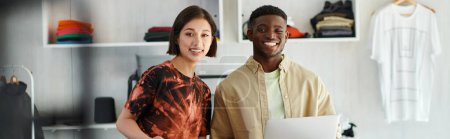 happy asian woman and african american man with laptop smiling at camera in print studio, banner
