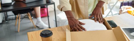 Téléchargez les photos : Vue recadrée de l'homme afro-américain emballant des vêtements dans une boîte en carton près d'une tasse en papier dans un atelier, bannière - en image libre de droit
