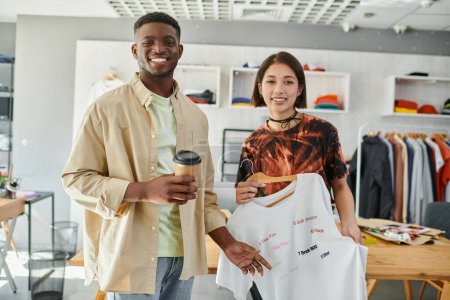 Foto de Diseñadores de moda multiétnicos felices con camiseta de moda y taza de papel mirando a la cámara en el taller - Imagen libre de derechos