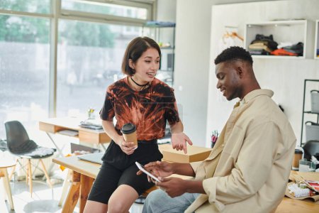Foto de Asiático diseñador con papel taza hablando con africano americano colega con digital tablet en atelier - Imagen libre de derechos