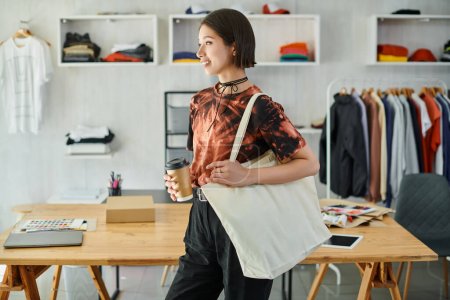 fröhliche und stilvolle asiatische Frau mit Leineneinkaufstasche und Getränk zum Mitnehmen in modernem Druckstudio