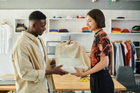 smiling asian designer giving canvas shopping bag to african american colleague in print studio