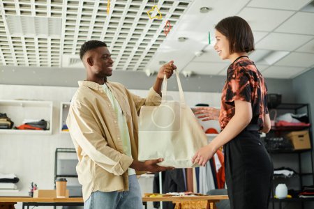 glücklicher afrikanisch-amerikanischer Designer mit einer Einkaufstasche aus Leinwand in der Nähe eines asiatischen Kollegen in einer Druckwerkstatt