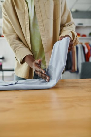 Foto de Recortado vista de africano americano trabajador de textil impresión estudio plegable ropa prolijamente en mesa - Imagen libre de derechos