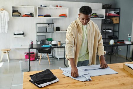 junger und fleißiger afrikanisch-amerikanischer Mann faltet Kleidungsstücke auf dem Tisch im Druckereistudio, Kleinunternehmen