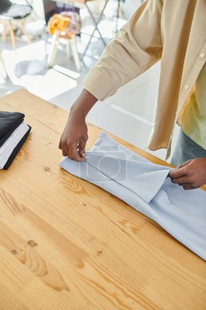 partial view of young skilled african american man folding clothes carefully in modern print studio