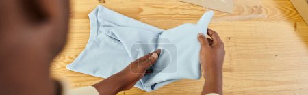 vista recortada del trabajador afroamericano calificado plegando la ropa en la mesa en atelier de impresión, bandera