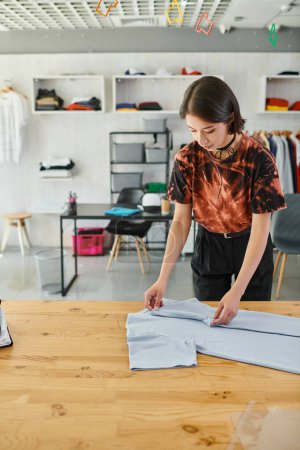 Foto de Joven y elegante mujer asiática plegable ropa en la mesa en taller de impresión, la industria de la moda - Imagen libre de derechos