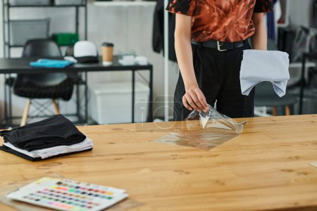 Foto de Vista recortada de la mujer joven embalaje doblado ropa en bolsa de plástico en la mesa en taller de impresión - Imagen libre de derechos
