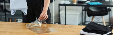 Foto de Vista recortada de la mujer embalaje cuidadosamente doblado ropa en bolsa de plástico en la mesa en el atelier de impresión, bandera - Imagen libre de derechos