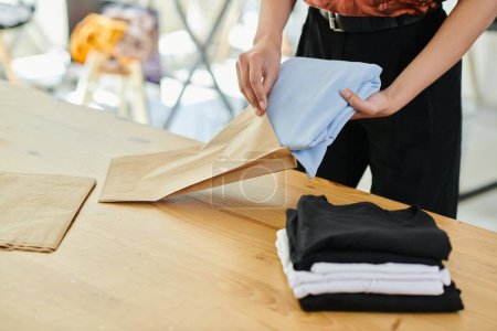 Foto de Vista recortada de mujer calificada embalaje doblado ropa en bolsa de papel en el estudio de impresión, pequeña empresa - Imagen libre de derechos
