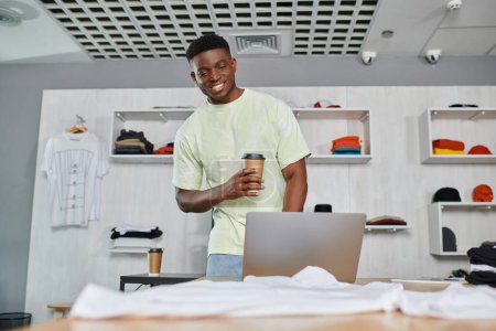 Foto de Sonriente diseñador afroamericano con café para ir a buscar portátil cerca de la ropa en el estudio de impresión - Imagen libre de derechos