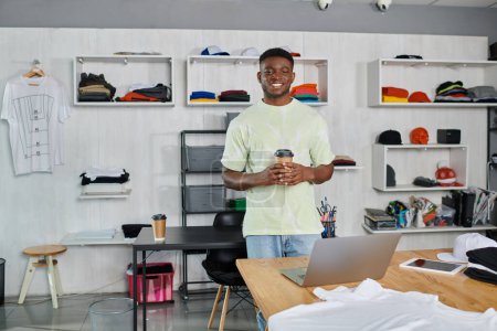 Foto de Diseñador afroamericano feliz con café para ir mirando la cámara cerca del ordenador portátil en el taller de impresión - Imagen libre de derechos