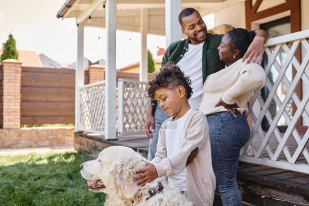 cheerful african american kid playing with dog near parents on backyard of their house, quality time