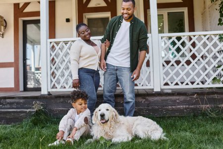 Foto de Tiempo familiar, feliz afroamericano padres mirando hijo jugando con perro en patio trasero de la casa - Imagen libre de derechos