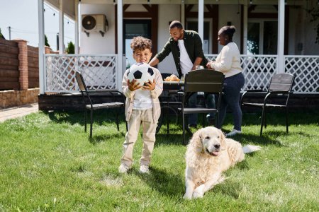 Foto de Tiempo familiar, niño afroamericano feliz celebración de fútbol cerca de los padres y el perro en el patio trasero de la casa - Imagen libre de derechos