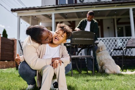 Foto de Africano americano madre besos mejilla de excitado hijo cerca marido y familia perro en backyard - Imagen libre de derechos