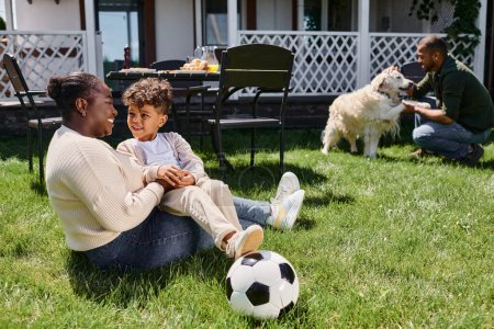 Foto de Feliz africano americano madre sentado en el césped con hijo al lado de fútbol, marido y perro en el patio trasero - Imagen libre de derechos
