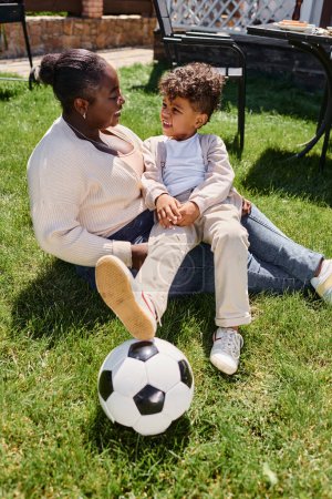 Foto de Feliz africano americano madre sentado en el césped con hijo al lado de fútbol pelota en patio trasero de la casa - Imagen libre de derechos