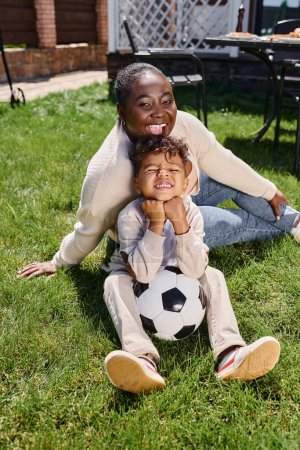 Foto de Alegre africano americano madre sentado en césped con hijo al lado de fútbol pelota en patio trasero de casa - Imagen libre de derechos