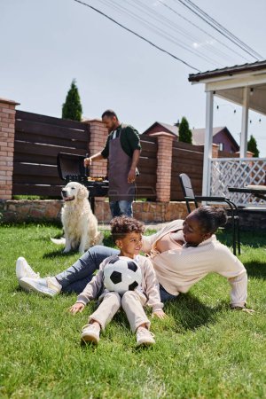 happy african american mother sitting on lawn with son near dog while husband cooking on bbq grill