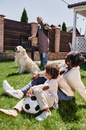 african american mother sitting on lawn with son near dog while happy husband cooking on bbq grill
