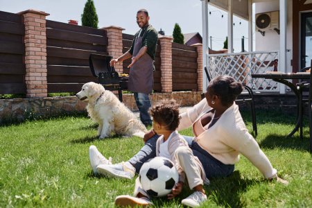 Afrikanisch-amerikanische Mutter sitzt mit Sohn neben Hund auf Rasen, während positiver Mann auf Grill kocht