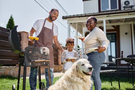 happy african american man preparing grilled corn on bbq grill near dog, wife and son on backyard