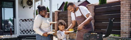 happy african american man serving grilled corn on plate of son near wife during bbq party, banner