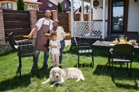 famille afro-américaine positive tenant des assiettes avec du maïs grillé près du barbecue grill et chien sur la cour