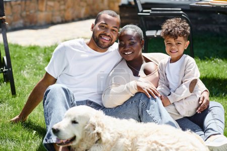 retrato familiar de la familia afroamericana positiva mirando a la cámara y sentado en el césped cerca del perro