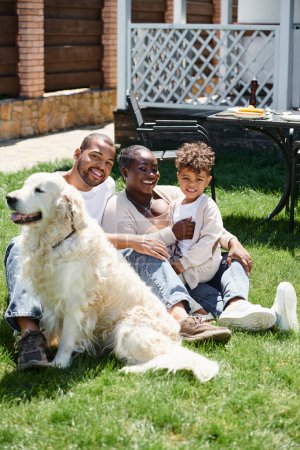 retrato familiar de alegres padres afroamericanos y su hijo sonriendo y sentado en el césped cerca del perro
