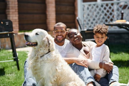 family portrait of joyful african american parents and son smiling and sitting on grass near dog