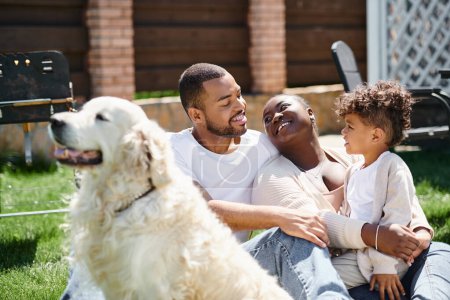 family time of joyful african american parents and son smiling and sitting on grass near dog