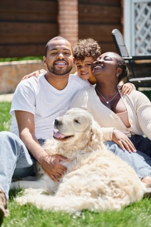 family portrait of cheerful african american parents and son smiling and sitting on lawn near dog