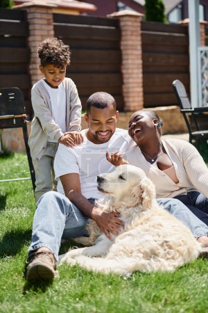 retrato de alegres padres afroamericanos y su hijo sonriendo y sentado en el césped verde cerca del perro