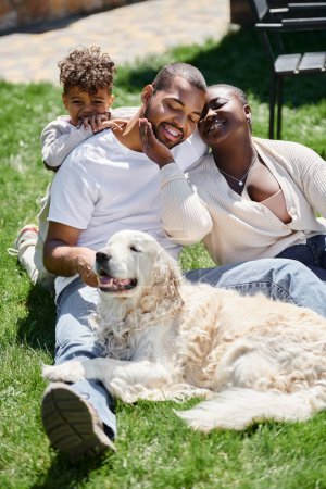 portrait of happy african american parents and son smiling and sitting on green lawn near dog