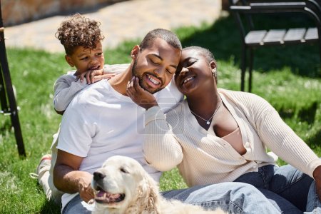 Familienmoment glücklicher afrikanisch-amerikanischer Eltern und Sohn lächelnd auf grünem Rasen neben Hund sitzend