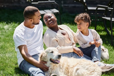 Familienmomente fröhlich afrikanisch-amerikanischer Eltern und Sohn lächelnd auf grünem Rasen neben Hund sitzend