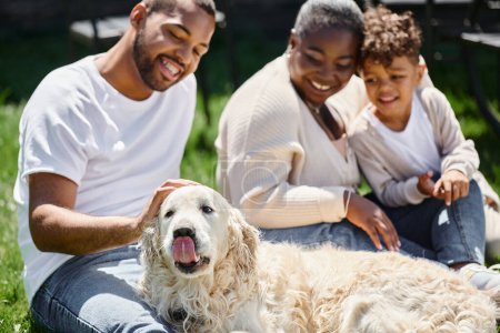 Foto de Momentos familiares de alegres padres afroamericanos e hijo sonriendo y sentado en el césped y acariciando perro - Imagen libre de derechos