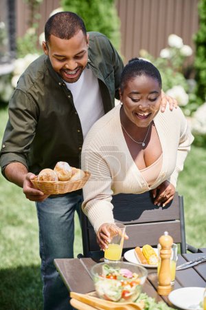 Foto de Feliz africano americano casado pareja sirviendo mesa en jardín, bollos y vaso de jugo de naranja - Imagen libre de derechos