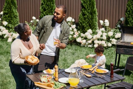 Téléchargez les photos : Heureux afro-américains parents servir table dans le jardin près de fils jouer avec chien sur arrière-cour - en image libre de droit