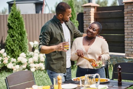 Afrikanisch-amerikanischer Mann umarmt glückliche Frau, die Salat in Glasschüssel mixt, während sie BBQ im Hinterhof hat