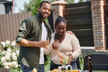 joyful african american man hugging wife mixing salad in glass bowl while having bbq on backyard